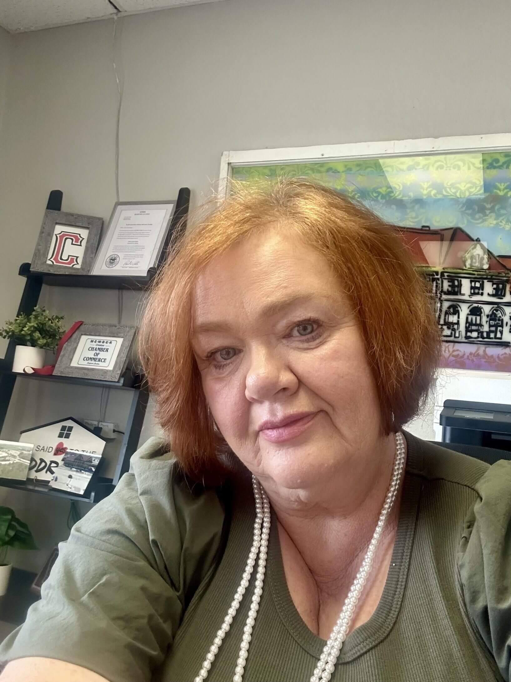 Smiling woman with short red hair wearing a green top and pearl necklace, seated in an office with shelves holding framed certificates and décor in the background.